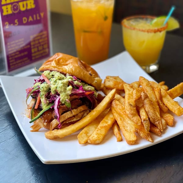 Yo Mommas Cantina's chicken sandwich with guacamole, slaw, fries, and two drinks on a dark table.