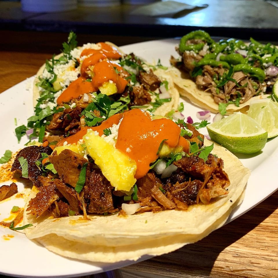 Three vibrant Mexican tacos on a white plate, two with red sauce and pineapple, one with green sauce.