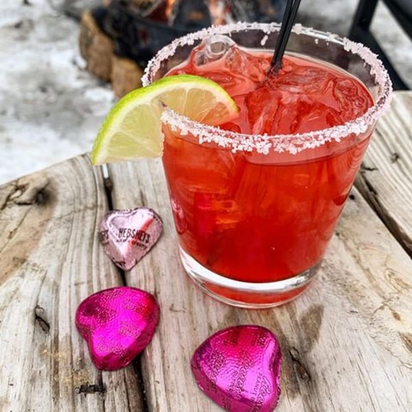 Red margarita with a lime wedge and salted rim, next to heart-shaped chocolates on a wooden surface.