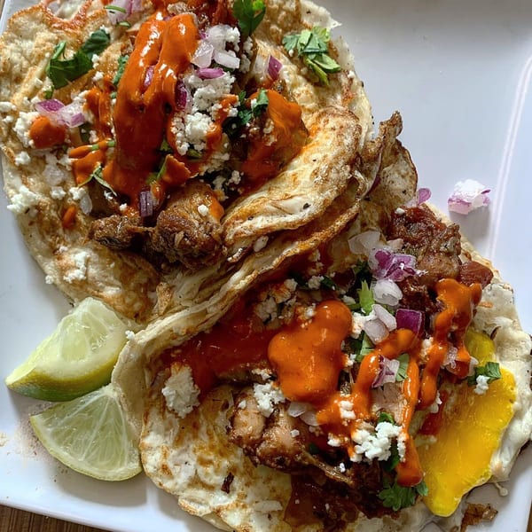 Two Mexican tacos with meat, cheese, red sauce, cilantro, and lime wedges on a white plate.