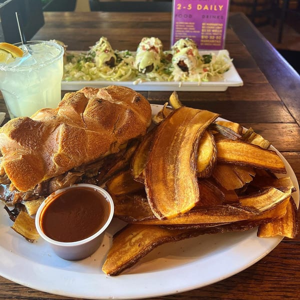 A large sandwich with a side of plantain chips, a dipping sauce, and a margarita.